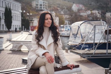 Kaygısız, bej elbiseli, sıcak ve rüzgarlı bir günde deniz manzarasının tadını çıkaran beyaz bir kadın. Bayan güzel bir kış gününün tadını çıkarıyor ve deniz havasını soluyor.