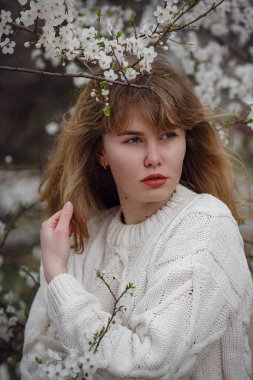 Bahar zamanı kiraz çiçeği bahçesinde beyaz süveter giyen güzel genç bir kadın. Açık hava moda fotoğrafı.