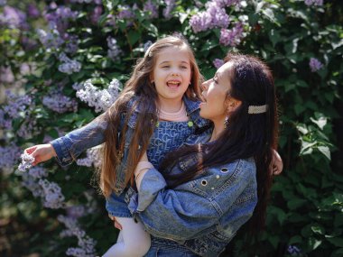 Kot elbiseli bir kadın bahar lilak bahçesinde eğleniyor. Anne ve çocuk gülümsüyor ve sarılıyorlar. Aile tatili ve birliktelik. Anneler Günün kutlu olsun.!