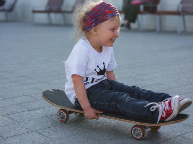 Küçük, güzel kız kaykay sürmeyi beyaz tişört ve kot pantolonla öğreniyor. Parkta açık hava sporları. Moda çocuğu konsepti