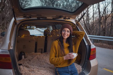 Arabanın bagajında oturan kadın. Gitmeye hazırlanıyorum. Arabasının bagajında oturan gülen genç bir kadın. Sonbahar araba gezisi. Hippi kurbağa seyahati, Sonbahar Kaçışı