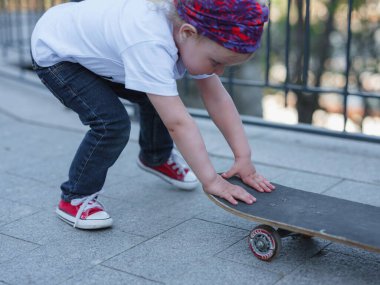 Küçük, güzel kız kaykay sürmeyi beyaz tişört ve kot pantolonla öğreniyor. Parkta açık hava sporları. Moda çocuğu konsepti