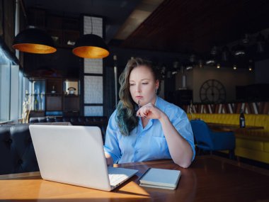 İşine odaklan. Kendine güveni tam genç bir kadın. Yaratıcı kafenin penceresinin yanında otururken dizüstü bilgisayarda çalışıyor.