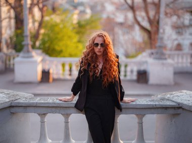 Siyah takım elbiseli kızıl genç kadın bahar şehrinde yürüyor. İş kadını iş gününden sonra gün batımının tadını çıkarıyor.