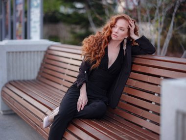 Kızıl genç kadın bahar şehrinde yürüyor. İş kadını bankta oturuyor.