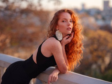 Siyah takım elbiseli kızıl genç kadın bahar şehrinde yürüyor. İş kadını iş gününden sonra gün batımının tadını çıkarıyor.