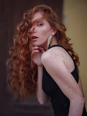 Siyah takım elbiseli kızıl genç kadın bahar şehrinde yürüyor. Moda trendi. Eski binanın yakınında poz veren şık bir genç bayan. El sallayan saç ve doğal makyaj..