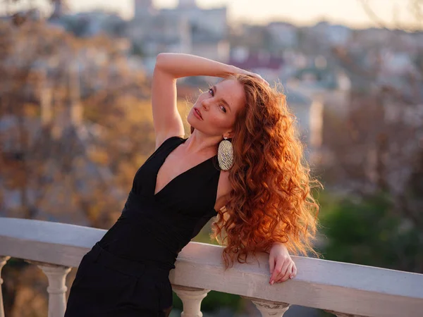 Siyah takım elbiseli kızıl genç kadın bahar şehrinde yürüyor. İş kadını iş gününden sonra gün batımının tadını çıkarıyor.