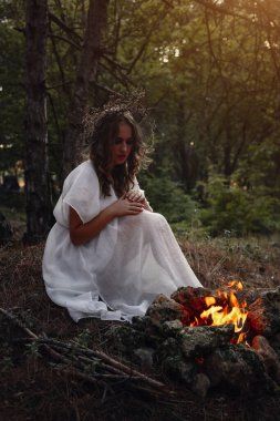 Sonbahar ormanında uzun beyaz elbiseli, gizemli gotik kadın. Karanlık fantezi yaratığı. Cadı ateşin yanında oturuyor.