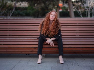 Kızıl genç kadın bahar şehrinde yürüyor. İş kadını bankta oturuyor.