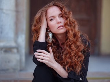 Siyah takım elbiseli kızıl genç kadın bahar şehrinde yürüyor. Moda trendi. Eski binanın yakınında poz veren şık bir genç bayan. El sallayan saç ve doğal makyaj..