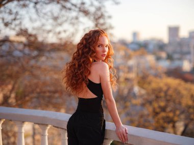 Siyah takım elbiseli kızıl genç kadın bahar şehrinde yürüyor. İş kadını iş gününden sonra gün batımının tadını çıkarıyor.