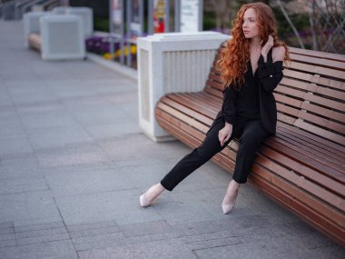 Kızıl genç kadın bahar şehrinde yürüyor. İş kadını bankta oturuyor.