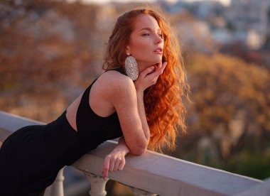 Siyah takım elbiseli kızıl genç kadın bahar şehrinde yürüyor. İş kadını iş gününden sonra gün batımının tadını çıkarıyor.