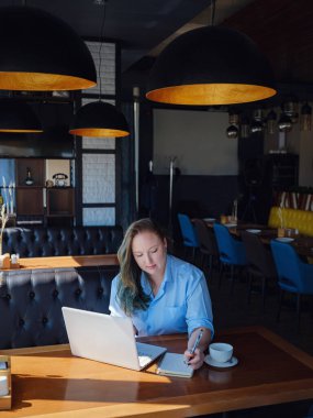 İşine odaklan. Kendine güveni tam genç bir kadın. Yaratıcı kafenin penceresinin yanında otururken dizüstü bilgisayarda çalışıyor.