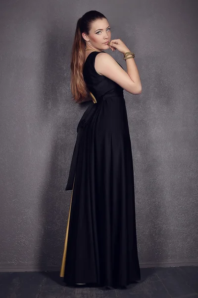 Young brunette lady in black ang golden dress posing on grey background