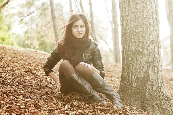 Menina sentada perto de uma árvore em um parque em um monte de folhas — Fotografia de Stock