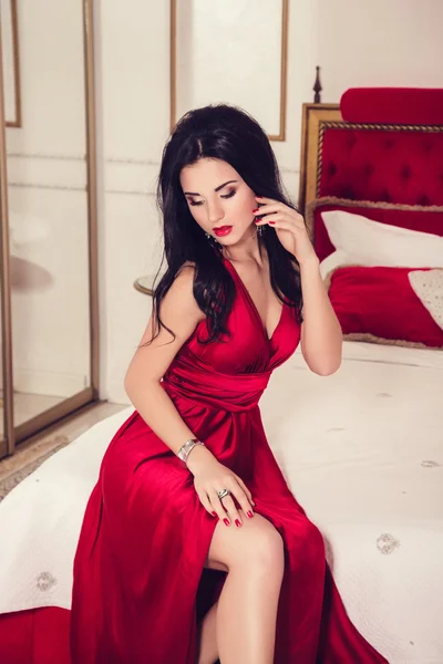 Αίγλη σέξι γυναίκα με μαύρα μαλλιά στο κομψό κόκκινο φόρεμα σε πολυτελή κρεβατοκάμαρα — Φωτογραφία Αρχείου