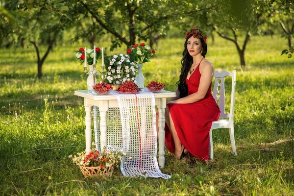 在一条长长的红裙子的女孩坐在花园里一张桌子 免版税图库图片