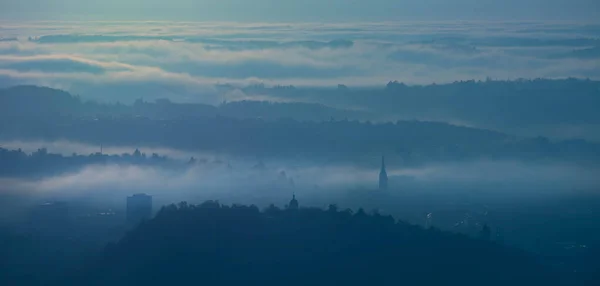 Amazing foggy sunrise over the city of Graz with Schlossberg hill and Church of the Sacred Heart of Jesus tower, in Styria region, Austria. Panoramic view from Plabutsch mountain on autumn morning.