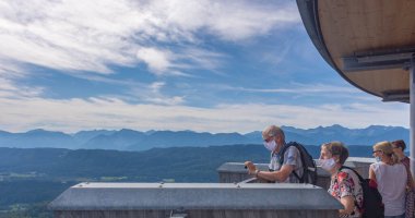 Klagenfurt, Avusturya - 26 Ağustos 2020: Coronavirus 'a karşı tıbbi maskeler takan insanlar, Worthersee Gölü' nün ve Piramidenkogel tahta kulesinin tepesinden dağların manzarasına hayran kaldılar.