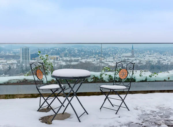 Table and chairs covered with snow on an empty terrace on Schlossberg hill, with beautiful view over the city of Graz,Styria region,Austria, in winter