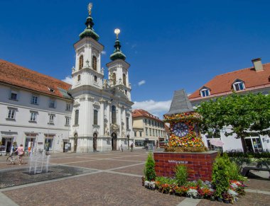 Graz, Avusturya-Temmuz 07, 2020: Ünlü saat kulesi, şehrin ana gösterilerinden biri, çiçekler ve Mariahilfer kilisesi ve güneşli bir günde meydan, Styria bölgesi, Avusturya