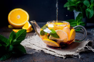 Portakal ve limon dilimli, böğürtlen, nane ve bal aromalı ev yapımı meyveli çay koyu kırsal arka planda. Sıcak sonbahar ya da kış içeceği. Sıcak su çaydanlığa dökülüyor..
