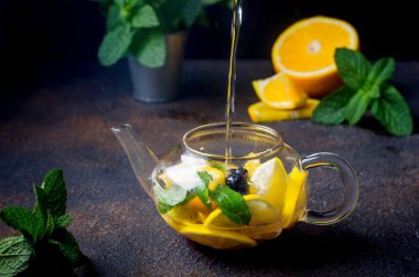 Portakal ve limon dilimli, böğürtlen, nane ve bal aromalı ev yapımı meyveli çay koyu kırsal arka planda. Sıcak sonbahar ya da kış içeceği. Sıcak su çaydanlığa dökülüyor..