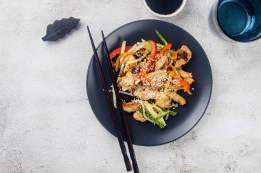 Çin yemek çubuklarıyla birlikte açık gri arka planda siyah bir kasede sebze tavası ve soslu tavuk eti ve susam. Geleneksel Asya yemekleri. 