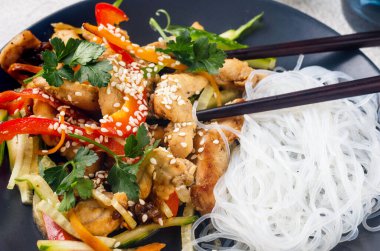 Sebzeli tavuk eti, Çin pilavı, erişte sosu ve açık gri arka planda Çin yemek çubukları olan siyah bir kasede susam. Geleneksel Asya yemekleri. 