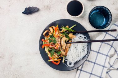 Sebzeli tavuk eti, Çin pilavı, erişte sosu ve açık gri arka planda Çin yemek çubukları olan siyah bir kasede susam. Geleneksel Asya yemekleri. 