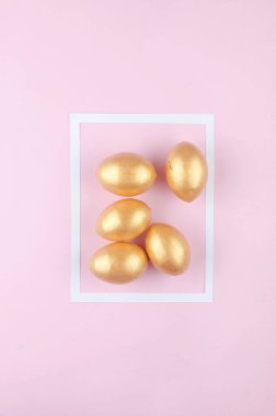 Pastel pembe arka planda beyaz çerçeveli altın renkli yumurtalar. Asgari paskalya konsepti. Mesaj için fotokopi alanı olan mutlu Paskalyalar. Üst görünüm, düz görünüm.