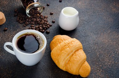 Taze kruvasan ve koyu beton arka planda bir fincan i sade kahve ve kahve çekirdeği. Sıcak içecekler, canlandırıcı bir kahvaltı..