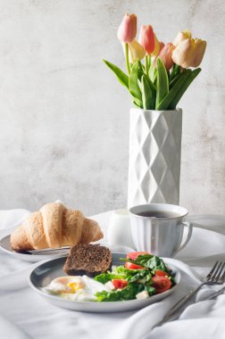 Sabah bahar sofrası dekorasyonu. Tabakta salata, yumurta, bir fincan kahve ve kruvasan, temiz beyaz masa örtüsünde vazoda taze laleler. Kahvaltı masası beyaz renkte ayarlanıyor.