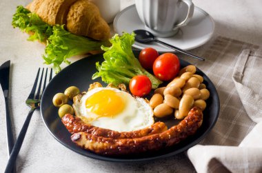 Geleneksel İngiliz kahvaltısı, kızarmış yumurta, sosis, fasulye, tabakta domates, kruvasan ve koyu arkaplanda kahve. Yukarıdan görüntüle 