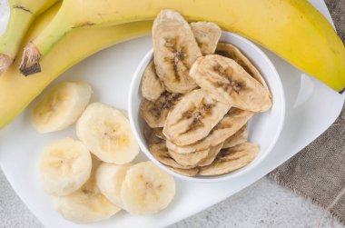 Olgun çiğ muz ve kurutulmuş muz dilimleri açık gri arka planda tabakta cipsler. Meyve cipsi. Sağlıklı beslenme konsepti, atıştırmalık, şekersiz. Üst görünüm, alanı kopyala.