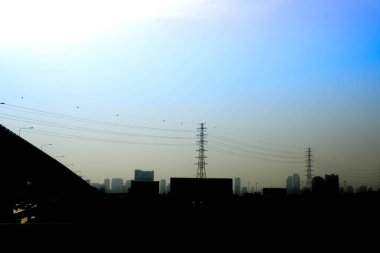 Elektrik iletim pilonu, modern kasabada mavi gökyüzüne ve binaya karşı yerleştirilmiş.