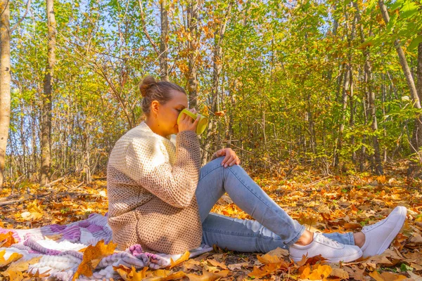 A woman in the autumn forest is drinking tea, sitting on a blanket, in a knitted sweater. Outdoor recreation
