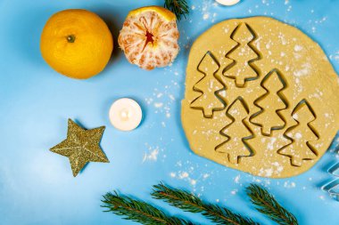 Çiğ hamur, köknar dalları, mandallar, mumlar ve mavi arka planda küçük hediyeler üzerinde köknar ağacı şeklinde zencefilli kurabiyeler. Yakın plan.