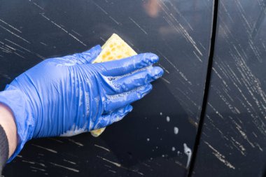 Mavi eldivenli bir kadının eli kirli bir arabanın kapıları boyunca köpükle kaplı sarı bir süngere öncülük eder. Yakın plan.