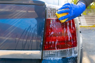 Mavi eldivenli bir kadın eli sarı köpüklü bir süngerle arabanın arka farını yıkar. Araba bakımı ve çamaşır.