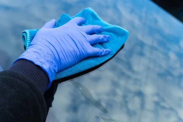 A woman's hand in a blue glove rubs the glass of a car in which the clouds are reflected. Car cleaning