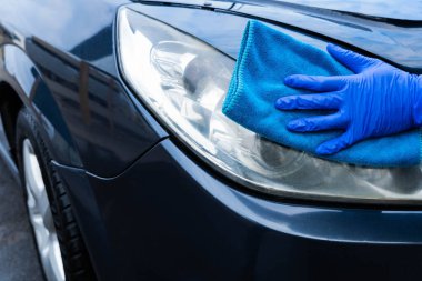 Mavi bir eldiven içinde mavi bir mikrofiber bez ile bir el bir arabanın farını ovar ve parlatır. Arabayı kendin yıka. Yakın plan.