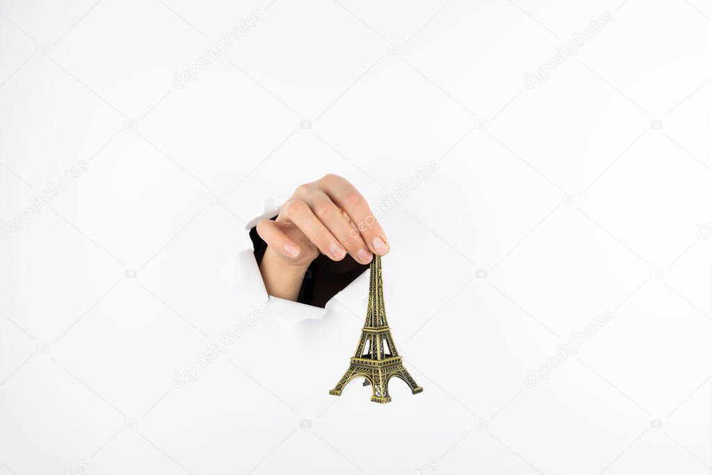 A female hand holds the eiffel tower statuette by the top on a white background. Hand goes through a hole in a white wall Free space for text