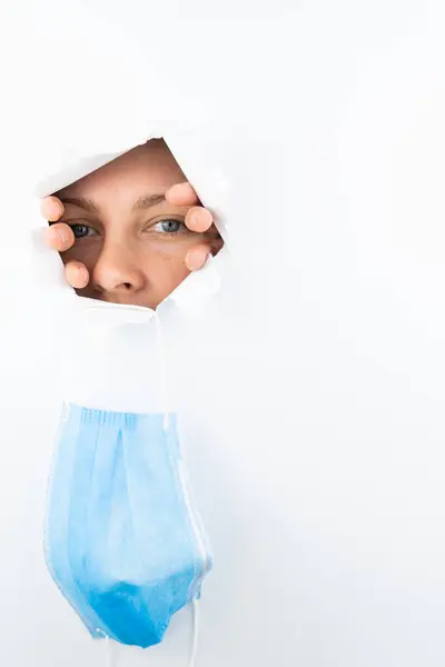 Девушка Голубыми Глазами Бросает Медицинскую Маску Через Дыру Белой Стене — стоковое фото