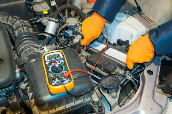 Изображение рук в оранжевых перчатках, измеряющих напряжение аккумулятора в автомобиле. Концепция ремонта и сервиса.