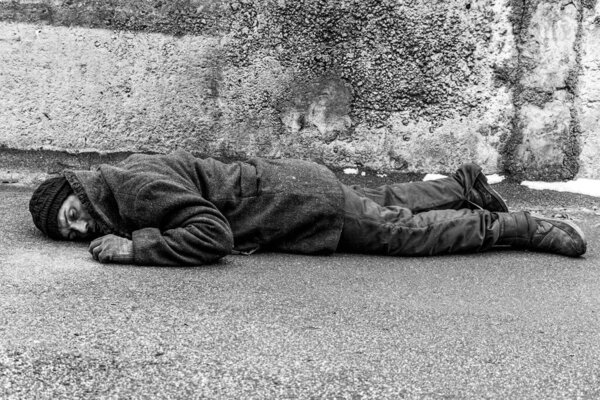 A dirty homeless man lies on the asphalt. A homeless man in warm clothes lies with his face to the ground.