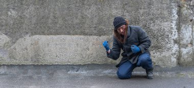 Zavallı, kirli adam, tek dizinin üzerine eğilmiş, yumruklarını gri duvara doğru gösteriyor. Metin için boş alan