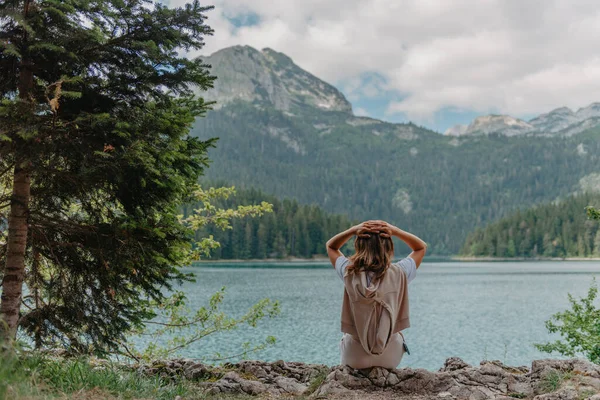 Mujer joven se sienta en la orilla del lago y mira al agua, vista trasera. Fin de semana en un hermoso gran lago. Turismo en Europa. Tranquilidad mental. Un hermoso lago en Montenegro. Lago en las montañas. — Foto de Stock